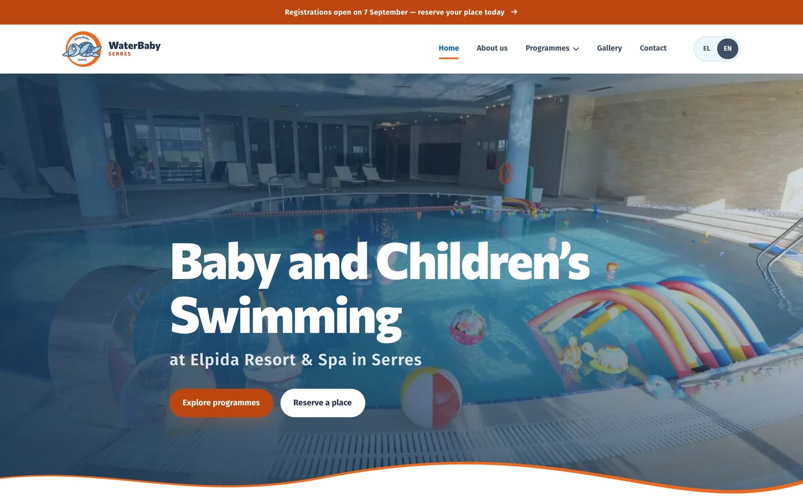 The WaterBaby home page: under an orange announcement bar and the navigation, with EN selected in the language switch, the headline “Baby and Children’s Swimming” and the line “at Elpida Resort & Spa in Serres” over a dimmed photograph of an indoor pool with inflatable rainbow arches, beach balls and floating toys, above an orange “Explore programmes” button, a white “Reserve a place” button and a wave edged in orange.