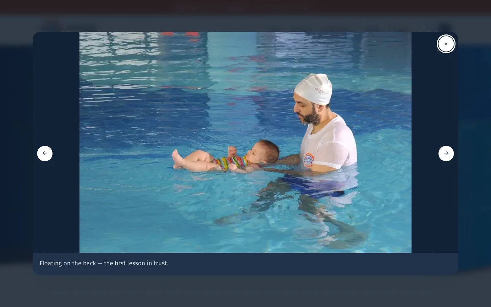 The lightbox open over the darkened gallery: a photograph of an adult in a white swim cap and a wet white T-shirt supporting a baby who floats on its back in the pool, the caption “Floating on the back — the first lesson in trust.” below it, round previous and next arrow buttons on either side, and a close button at the top right with a focus ring.
