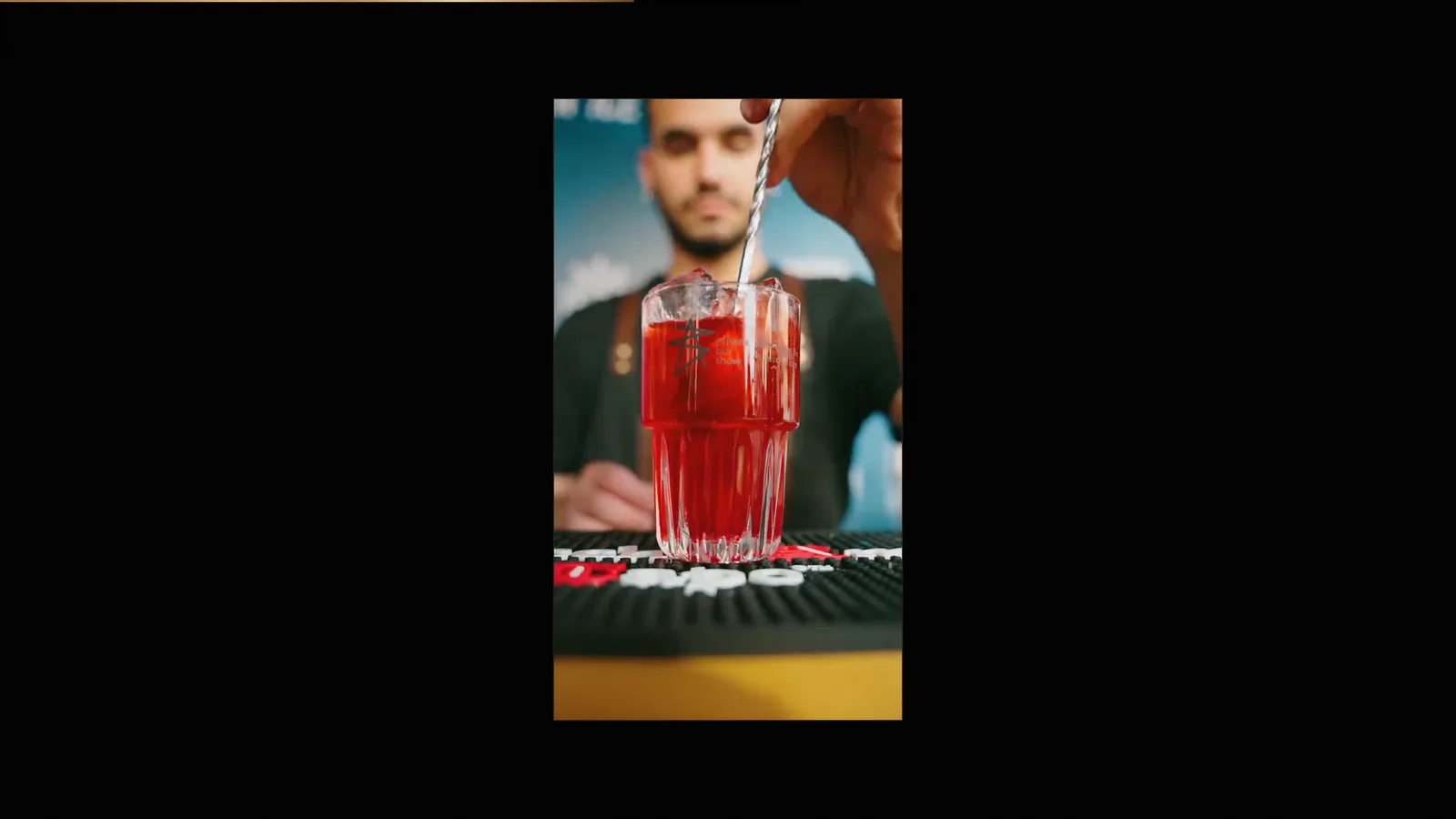 Portrait video frame of a bartender stirring a red cocktail, framed on a black page