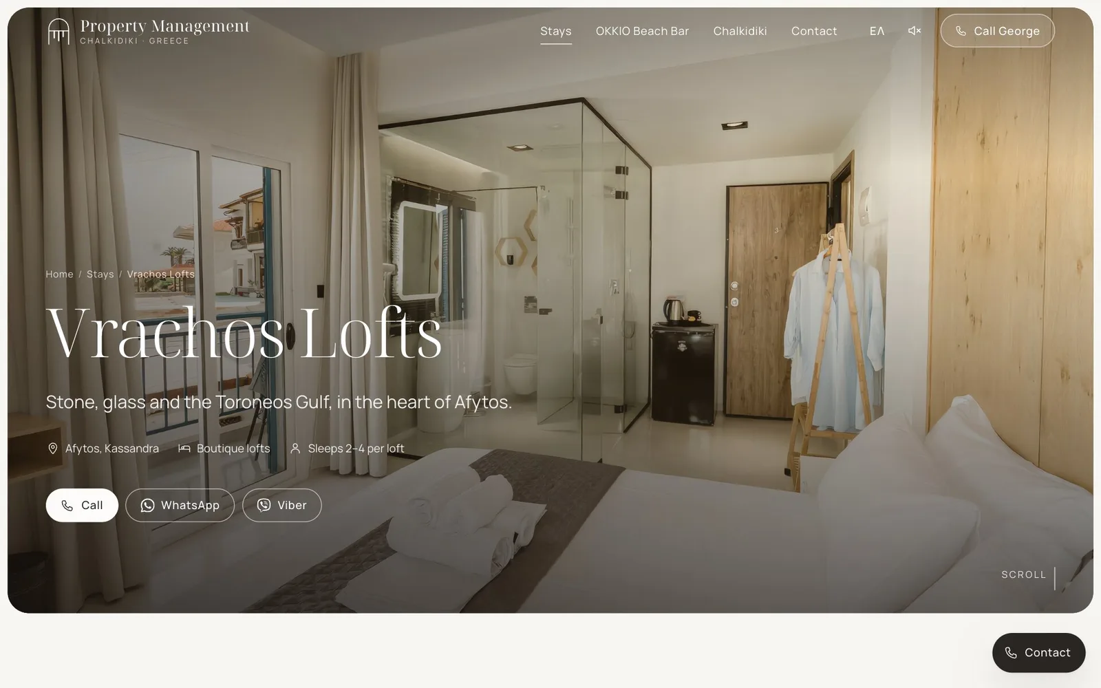 The Vrachos Lofts page: below the path Home / Stays / Vrachos Lofts, the title "Vrachos Lofts" and the line "Stone, glass and the Toroneos Gulf, in the heart of Afytos.", then the facts Afytos, Kassandra · Boutique lofts · Sleeps 2–4 per loft and Call, WhatsApp and Viber buttons, over a photograph of a loft with a glass-walled bathroom, an oak wall, a bed with rolled towels and a robe on a wooden stand.