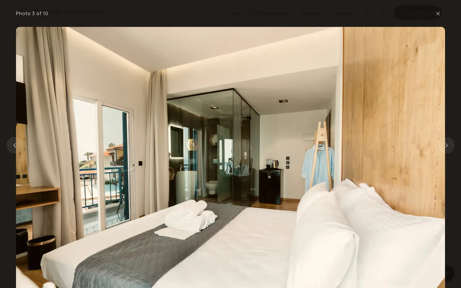 The photo viewer open over the Vrachos Lofts page, labelled "Photo 3 of 10", with a close button and previous and next arrows: a loft with a double bed, rolled towels, an oak wall, a glass shower enclosure and a balcony door.