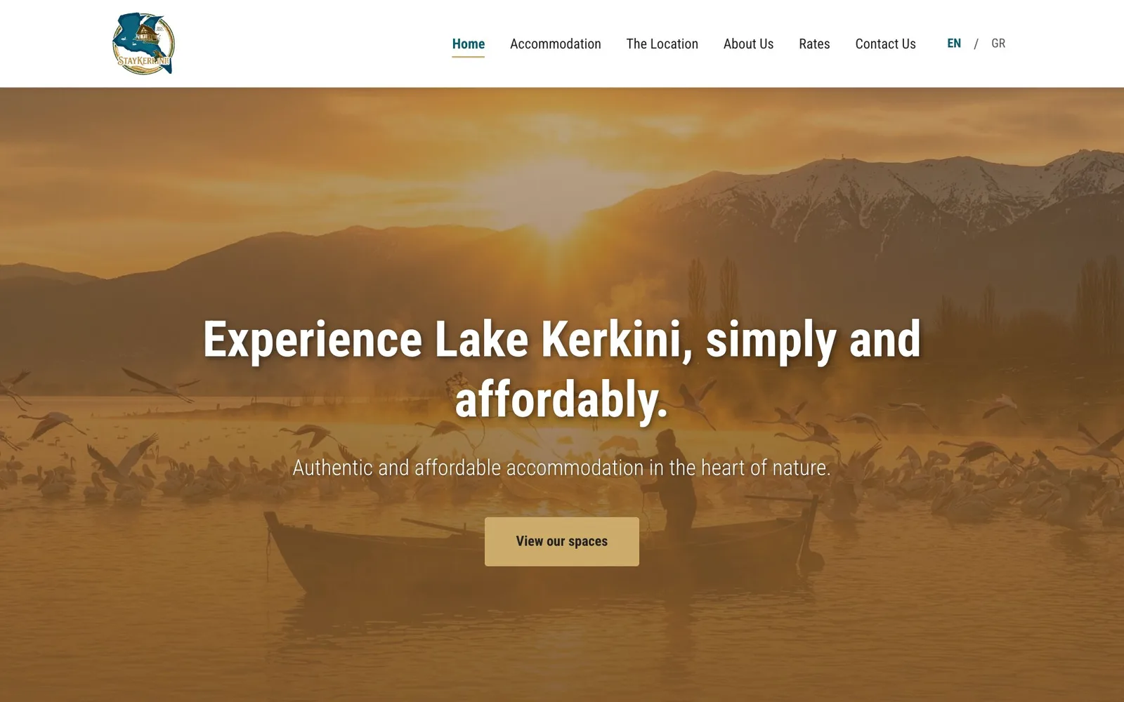 The StayKerkini home page: the round logo and the menu Home, Accommodation, The Location, About Us, Rates and Contact Us, with an EN / GR switch; over an amber-tinted photograph of Lake Kerkini with the sun low behind snow-capped mountains, a person in a wooden boat and pelicans and flamingos on the water and in the air, the headline "Experience Lake Kerkini, simply and affordably.", the line "Authentic and affordable accommodation in the heart of nature." and a gold "View our spaces" button.